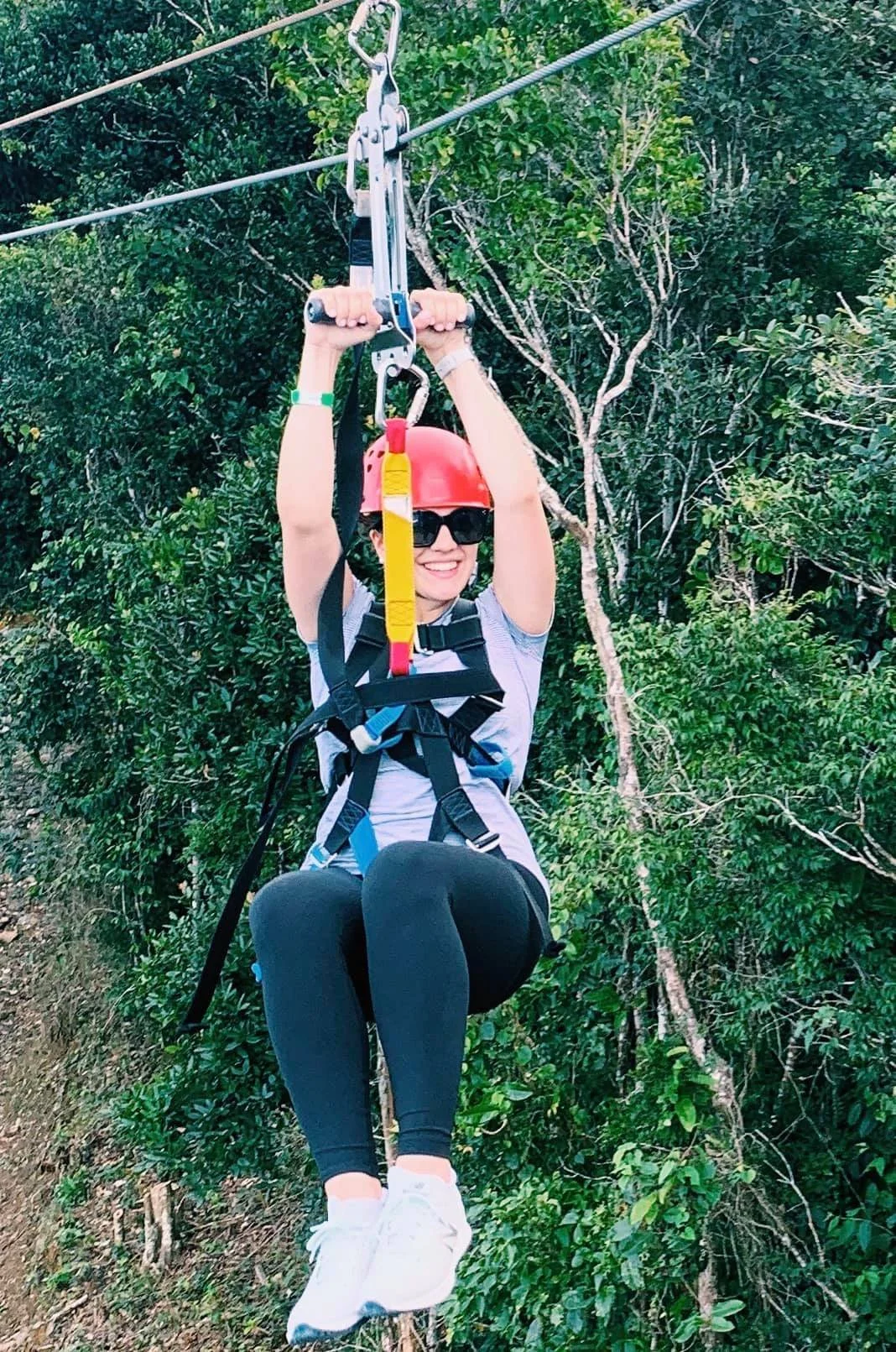 A woman wearing a red helmet and black sunglasses is zip-lining through a green forest. She is smiling and holding onto the zip line's handlebar, with trees in the background.