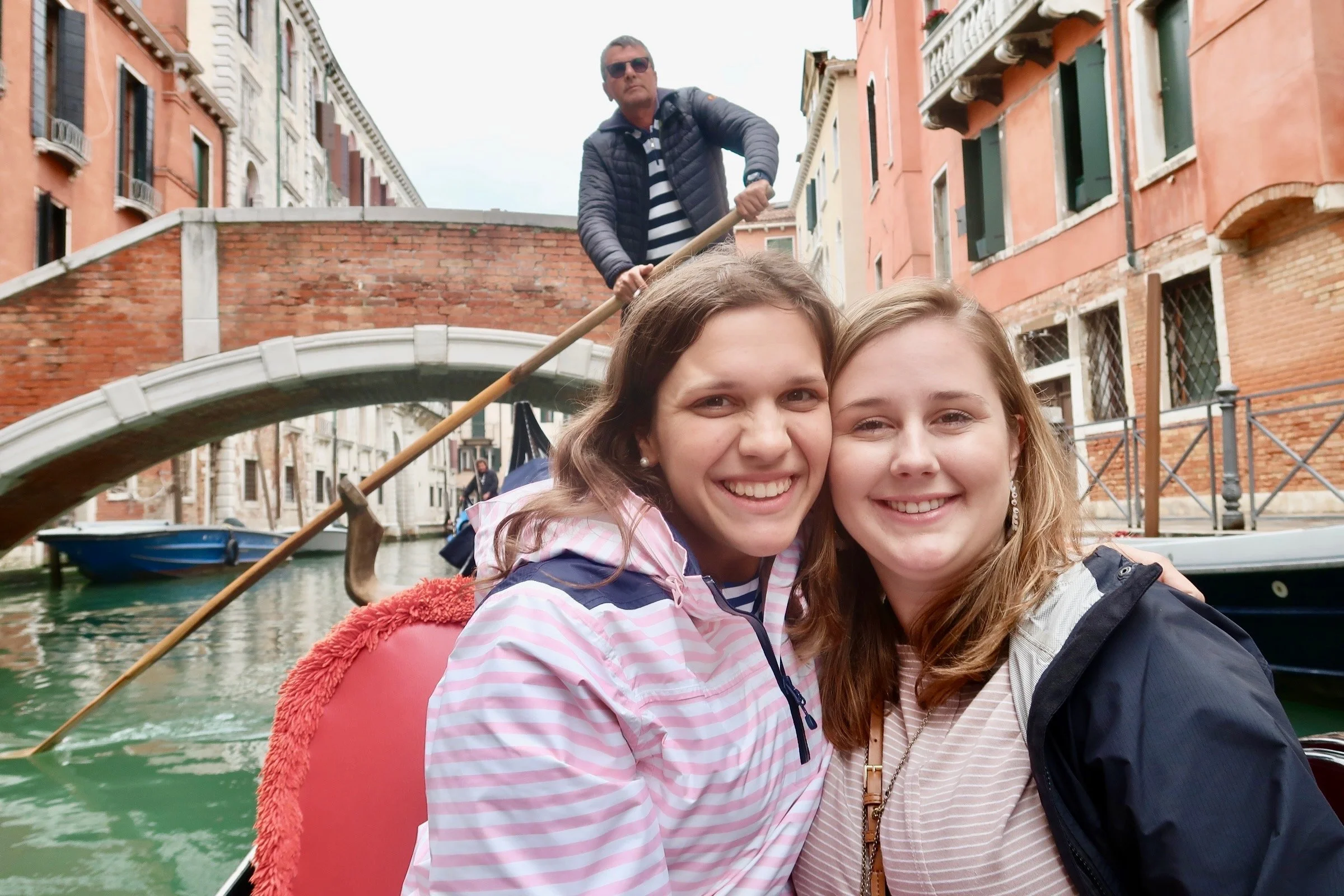 Two young women smiling for a selfie in a boat on a canal in Venice, Italy, with a gondolier standing on a bridge in the background.