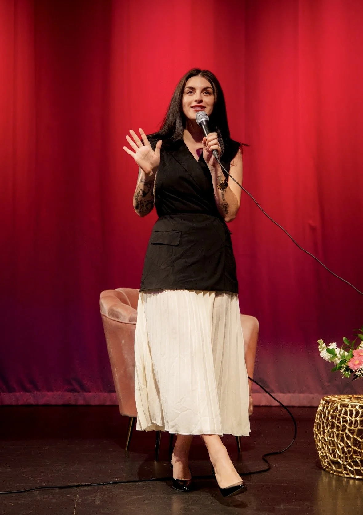 Woman with long dark hair, wearing a black sleeveless top and beige pleated skirt, speaking into a microphone on stage with a red curtain backdrop.