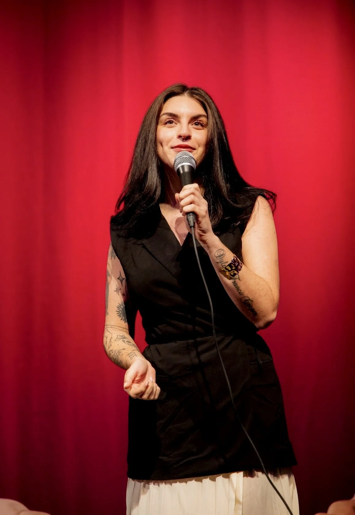 A woman with long dark hair holding a microphone on stage with a red curtain background.