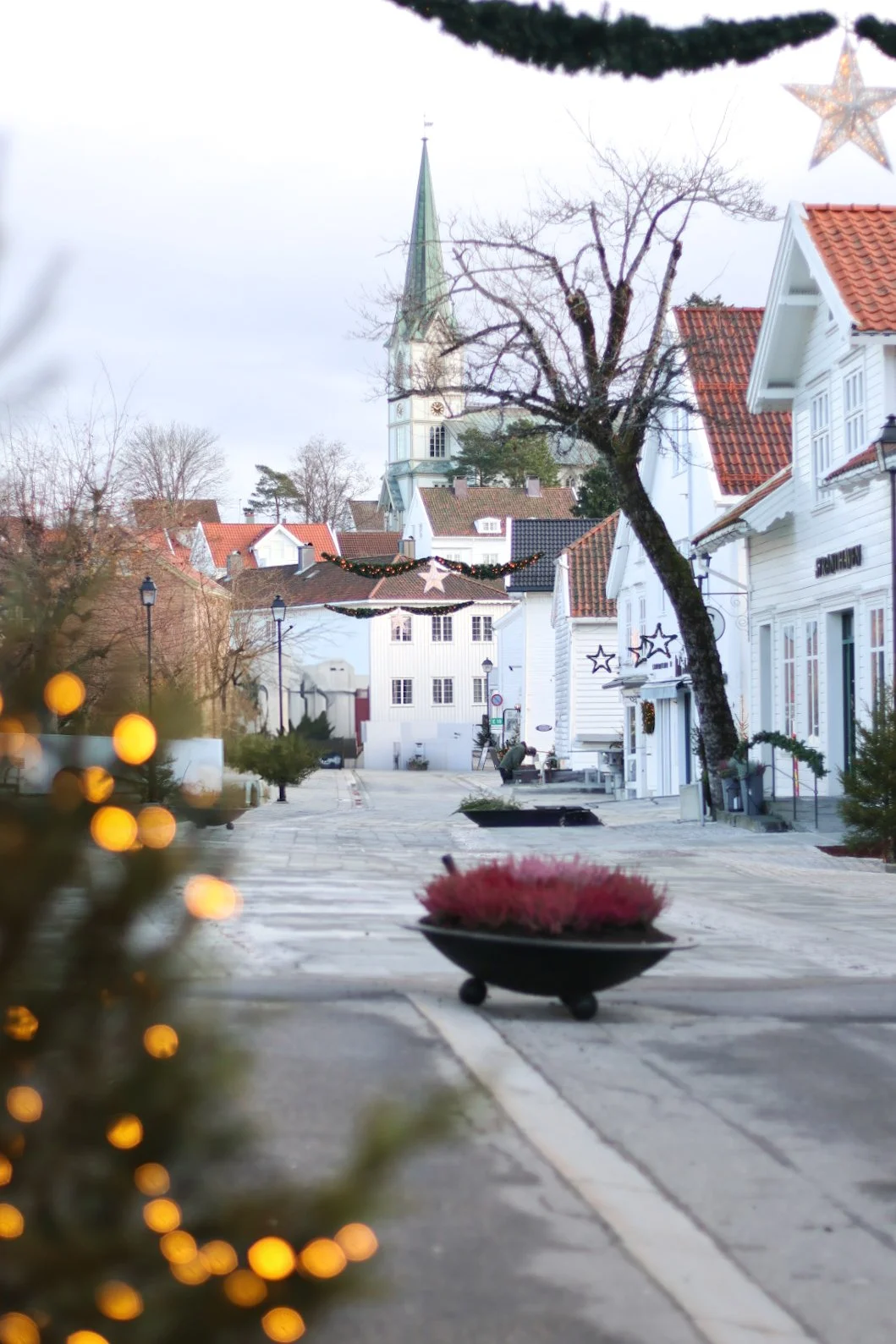 En bygate med juledekorasjoner, små tre med lys, julekuler, og en kirke i bakgrunnen. Gaten er tom og vinterlig.