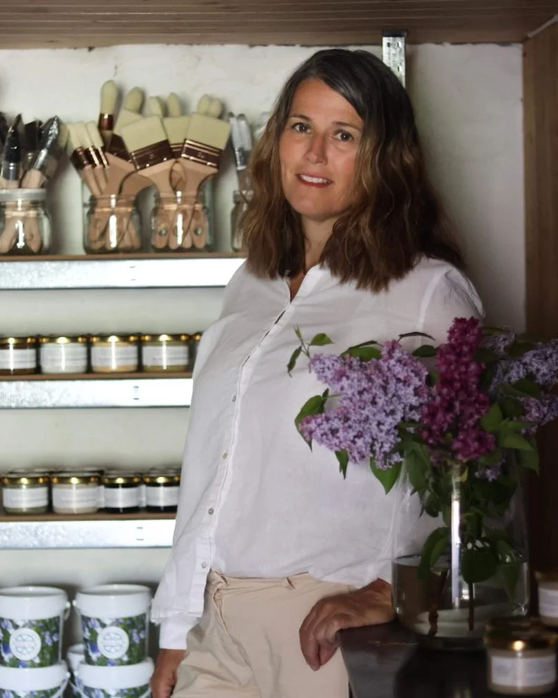 Kvinne med brunt, bølget hår står i et rom med hyller fylt med kosmetikk og produkter, og det er en vase med lilla og rosa blomster på et bord foran henne.