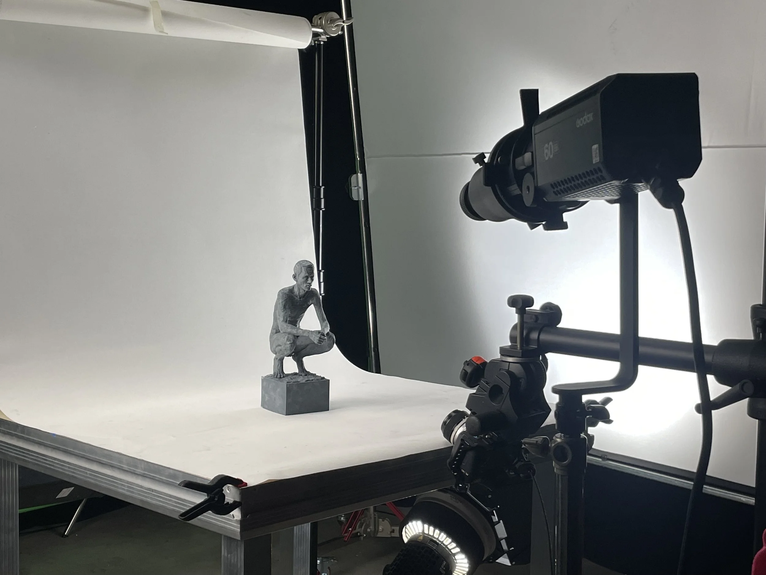 Une photographie d'un studio de production en cours, avec une sculpture de petite taille d'une personne assise en position accroupie, montée sur un socle, positionnée sur une table blanche. Un éclairage est disposé pour mettre en valeur la sculpture, avec un fond blanc et un équipement de tournage moderne.