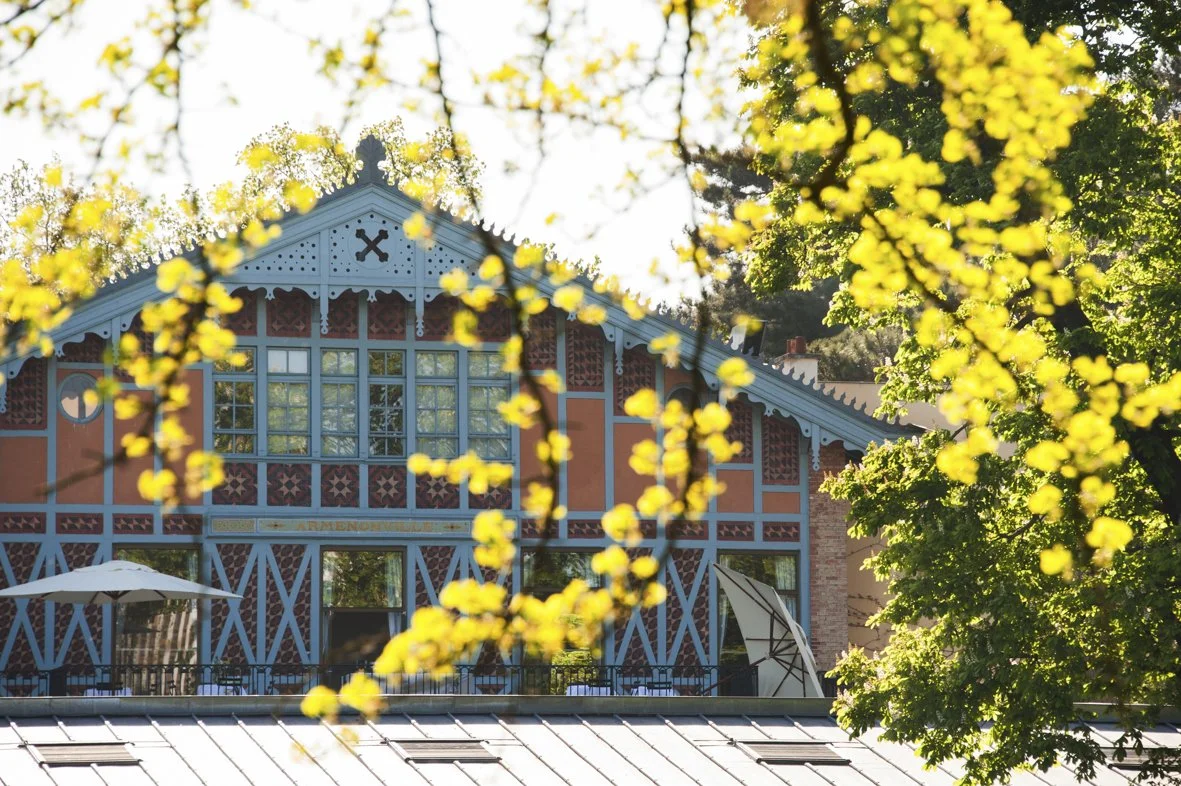 Une maison colorée avec une façade en bois peint en bleu et orange, entourée d'arbres avec des fleurs jaunes en premier plan