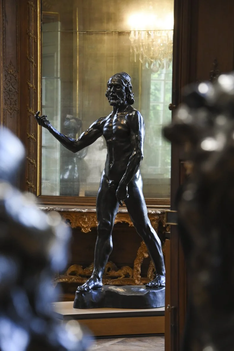 Statue en bronze d'un homme nu avec une barbe, tournant son corps légèrement vers la gauche, dans un intérieur avec un grand miroir et des meubles ornés.