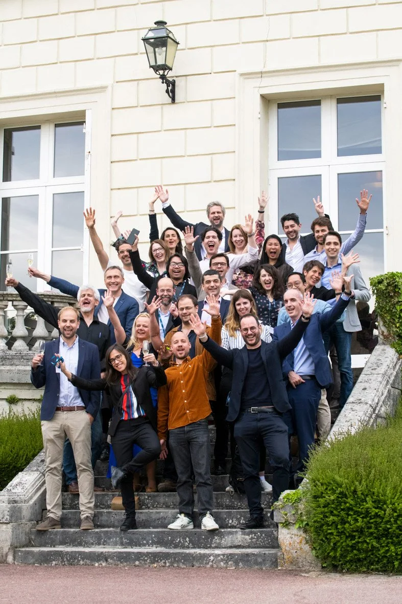 Un groupe de personnes souriantes et joyeuses posant sur des escaliers devant un bâtiment en pierre avec de grandes fenêtres, en train de célébrer.