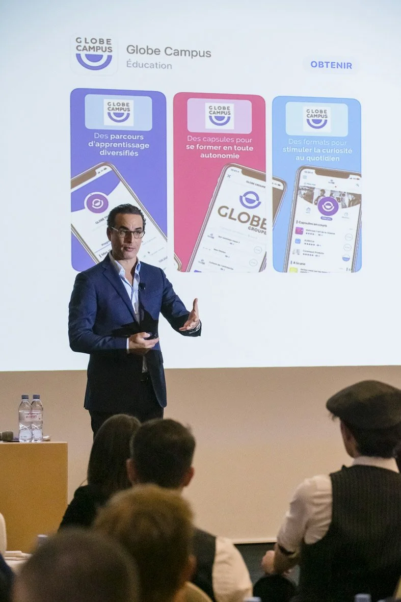 Un homme en costume bleu présente une diapositive sur une projection, expliquant des fonctionnalités de Globe Campus, avec un public assis.