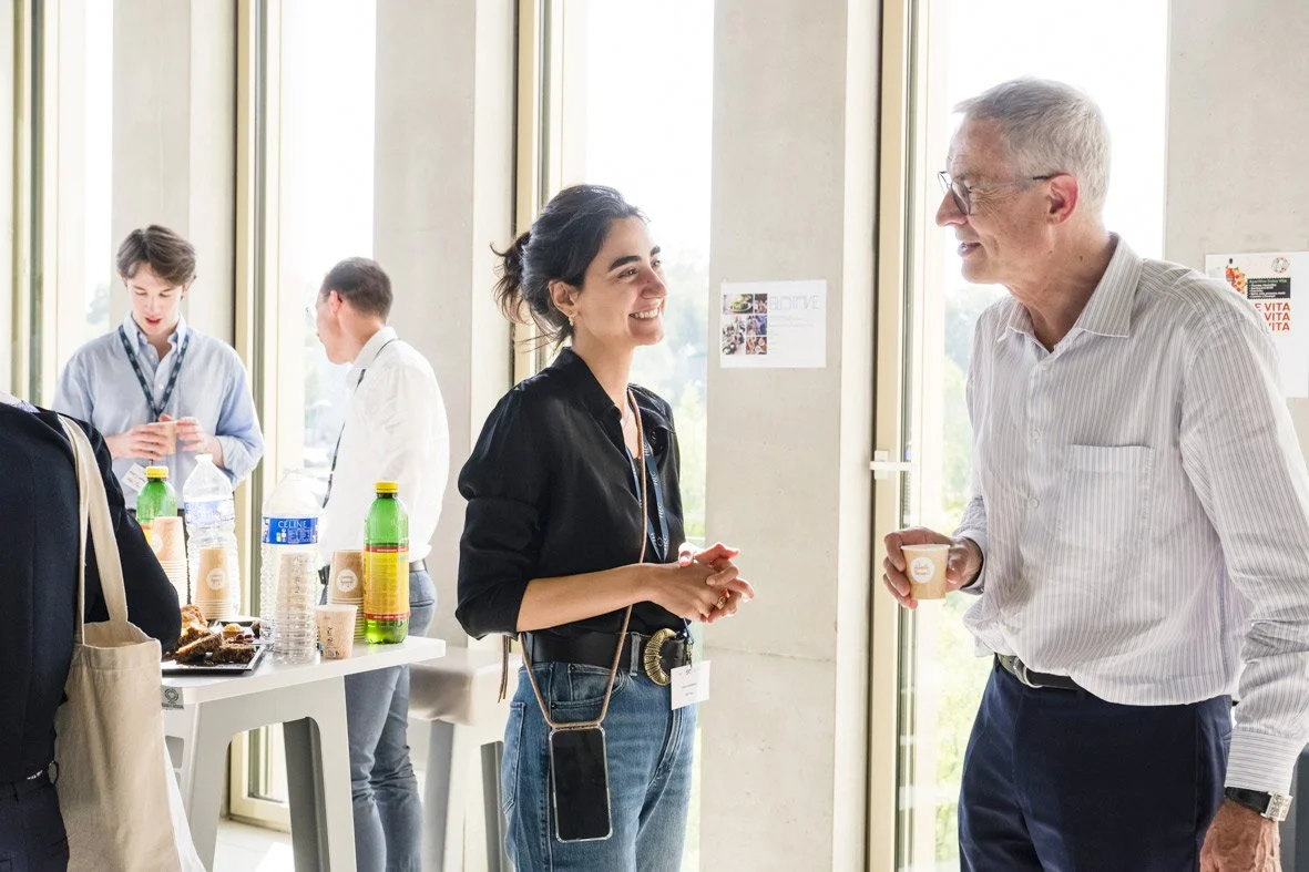 Deux personnes conversent joyeusement en tenant des tasses, dans un environnement lumineux avec de grandes fenêtres. Deux autres personnes sont au fond à gauche près d'une table avec des bouteilles et des snacks.
