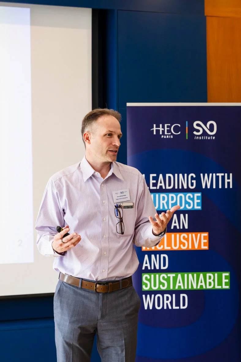 Un homme en chemise rayée donne une présentation lors d'une conférence, avec un panneau derrière affichant le logo de HEC Paris et des mots comme « Purpose », « Inclusive », « Sustainable ».