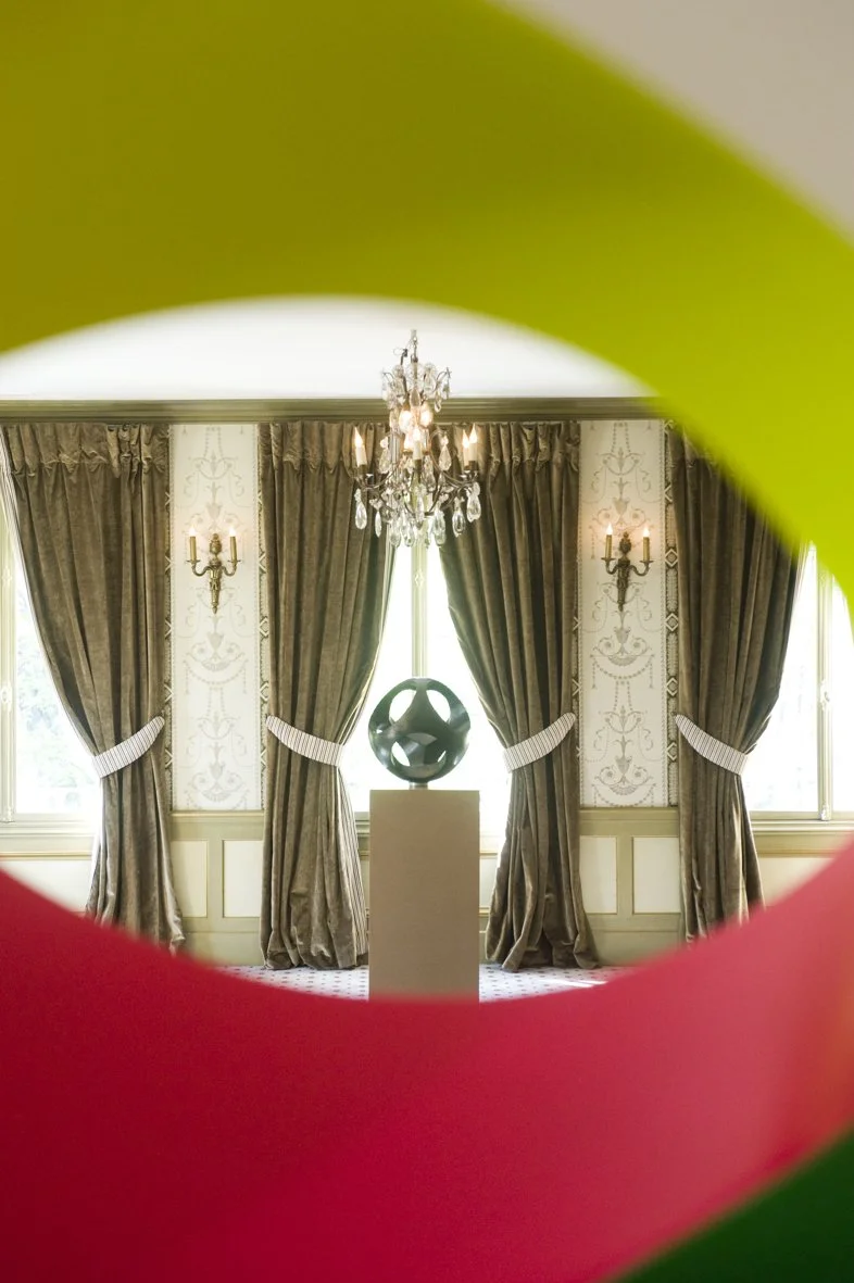 Une salle élégante avec des rideaux dorés, un chandelier et une sculpture noire en forme de cercle, vue à travers un objet coloré flou.