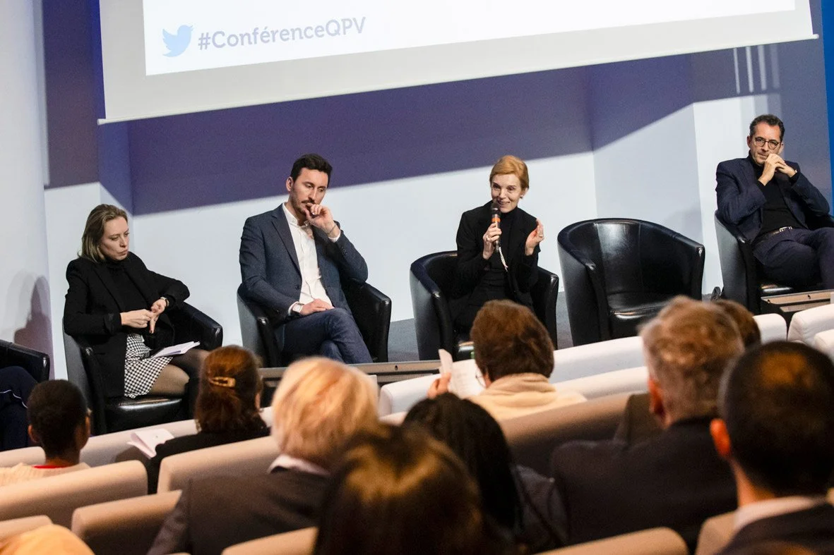 Une femme parle lors d'une table ronde ou d'un panel lors d'une conférence, avec un public en regard. Les autres membres du panel l'écoutent, assis dans des fauteuils noirs.