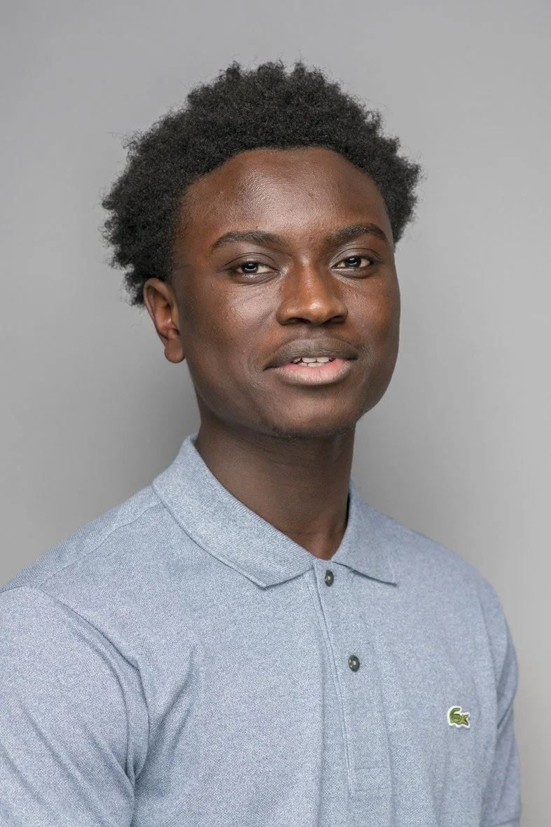 Un jeune homme noir avec des cheveux afro, portant un polo gris, regardant vers l'avant, avec un arrière-plan gris clair.