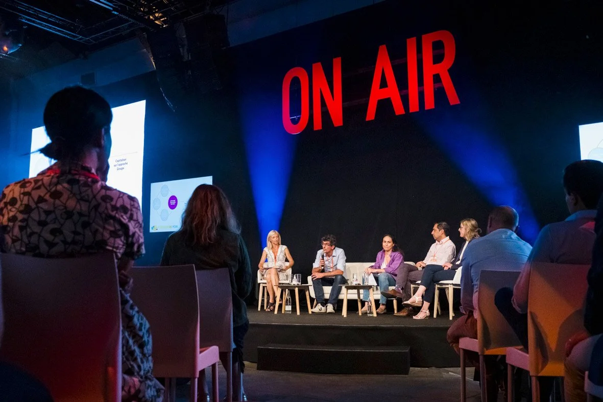 Une scène de conférence avec plusieurs intervenants assis sur scène, devant un grand panneau rouge avec l'inscription 'ON AIR'.