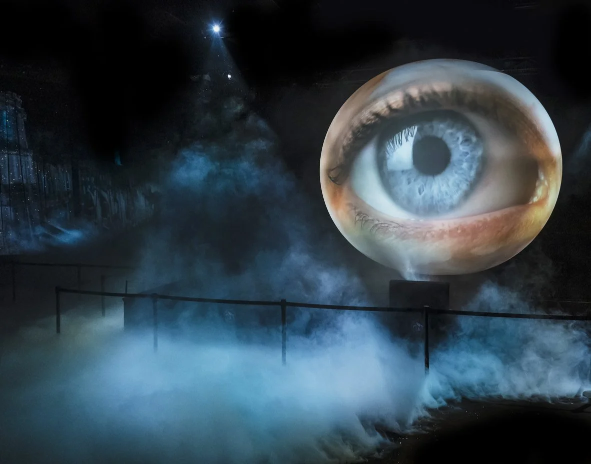 Une scène sombre avec de la brume et une projection d'un œil humain géant fixant vers l'avant, dans un environnement mystérieux ou dramatique.