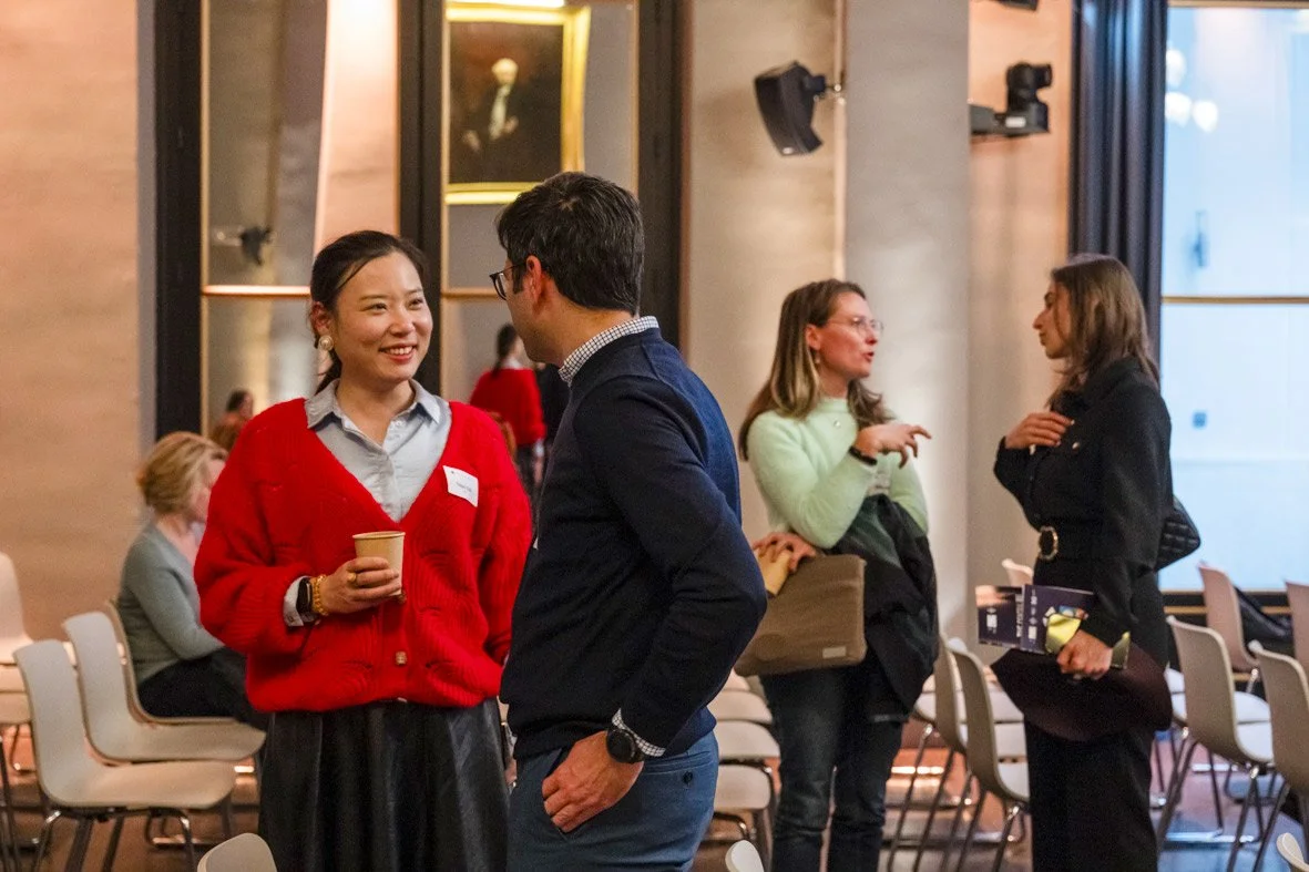 Quatre personnes discutent dans une salle lors d'un événement professionnel, deux d'entre elles en premier plan échangent un sourire, une tient une tasse en papier, toutes vêtues de vêtements décontractés.