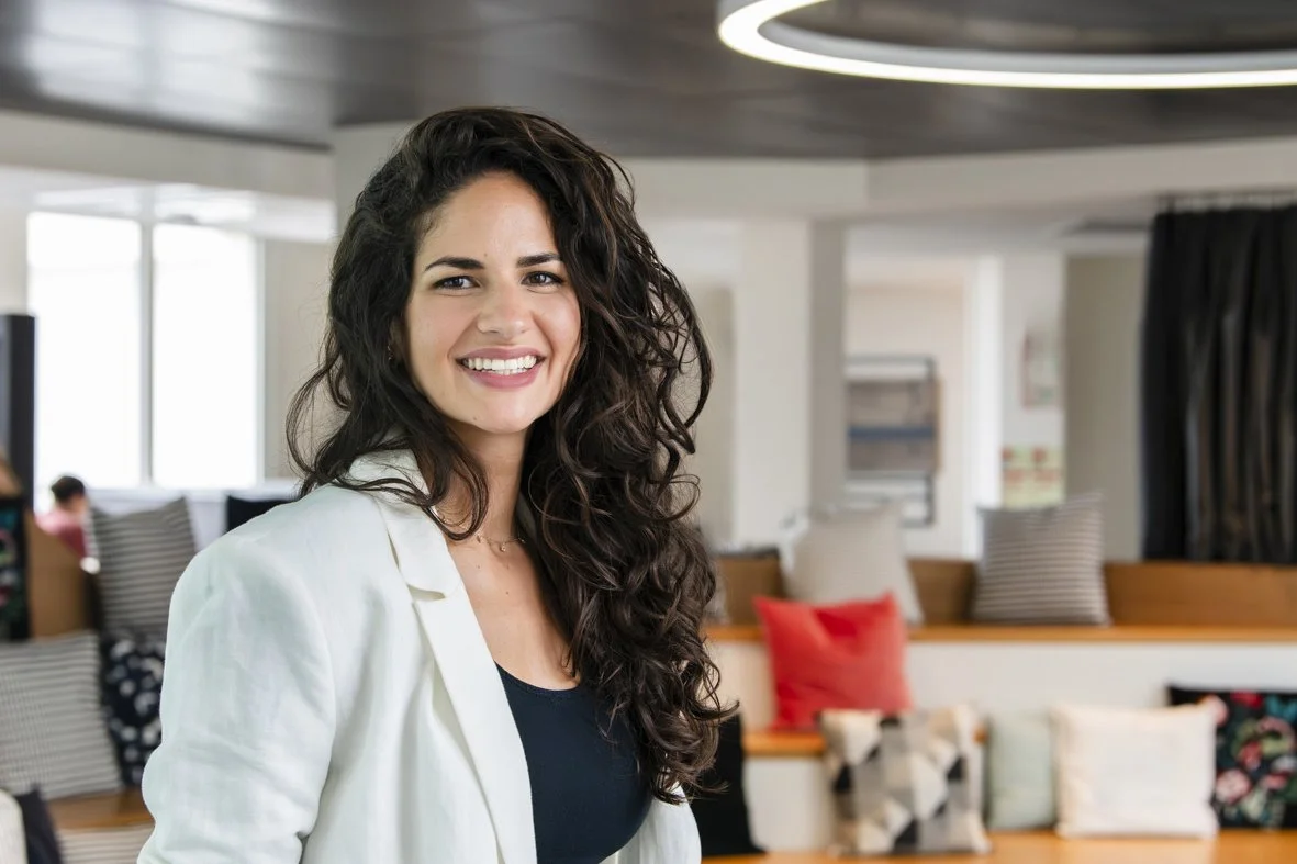 Une femme souriante avec des cheveux bouclés foncés, portant une veste blanche et un haut noir, dans un espace intérieur moderne avec des coussins colorés sur un banc.