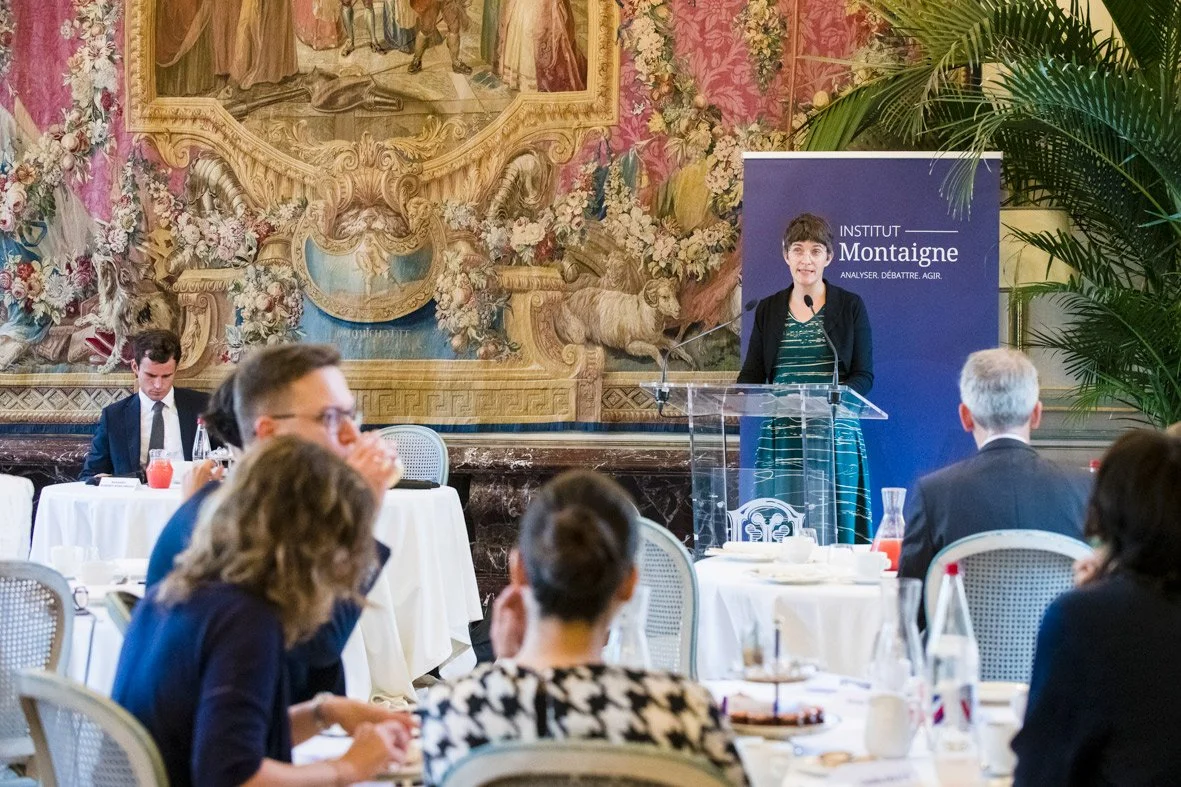 Une femme parle lors d'une conférence à l'Institut Montaigne, dans une salle décorée avec des murs recouverts de peintures anciennes et entourée de plantes vertes.