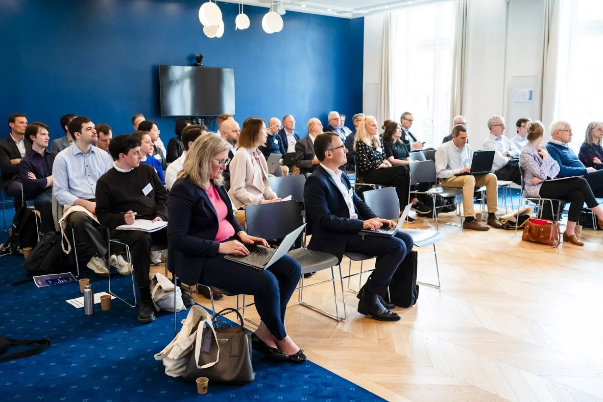 Une salle de conférence avec des personnes assises, certaines utilisant des ordinateurs portables, dans un environnement professionnel lumineux avec murs bleus et grands fenêtres.