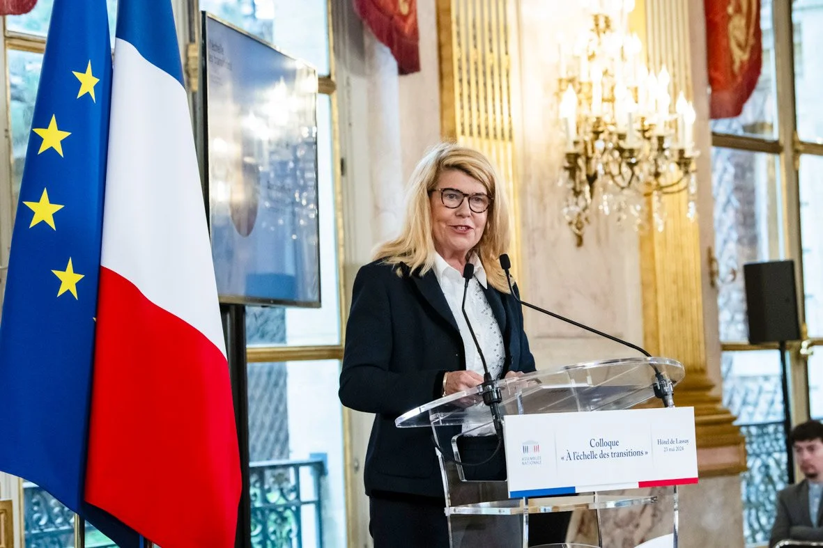 Une femme blonde avec des lunettes parle devant un micro à un podium lors d'une conférence dans une salle ornée de dorures et de grands lustres, avec le drapeau français et le drapeau de l'Union européenne à côté d'elle.