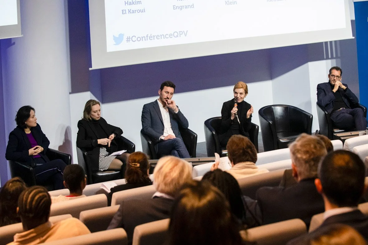Une conférence avec cinq intervenants assis sur scène face à une audience. La femme au centre parle en tenant un micro. Le fond affiche un écran avec du texte.