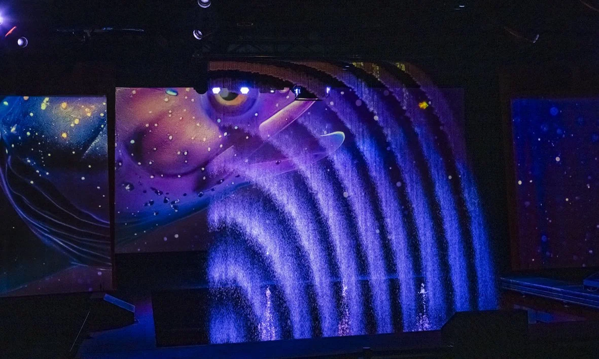 Une scène sombre avec un écran numérique affichant des motifs de galaxies, d'étoiles et d'eau en cascade en bleu et violet.