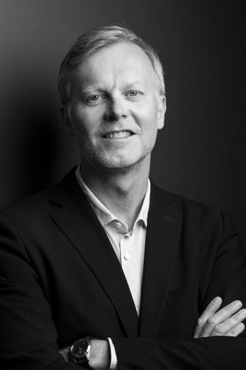 Un homme d'âge moyen, avec des cheveux gris et une barbe légère, porte un costume noir et une chemise blanche, pose avec les bras croisés, souriant, contre un fond sombre en noir et blanc.