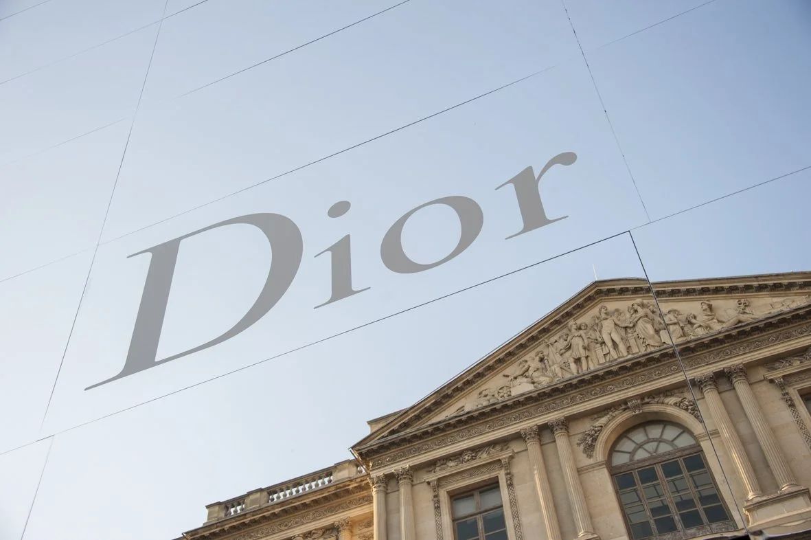 Une façade de bâtiment ancien avec une sculpture au-dessus de la porte et le logo Dior écrit en grand dans le ciel.