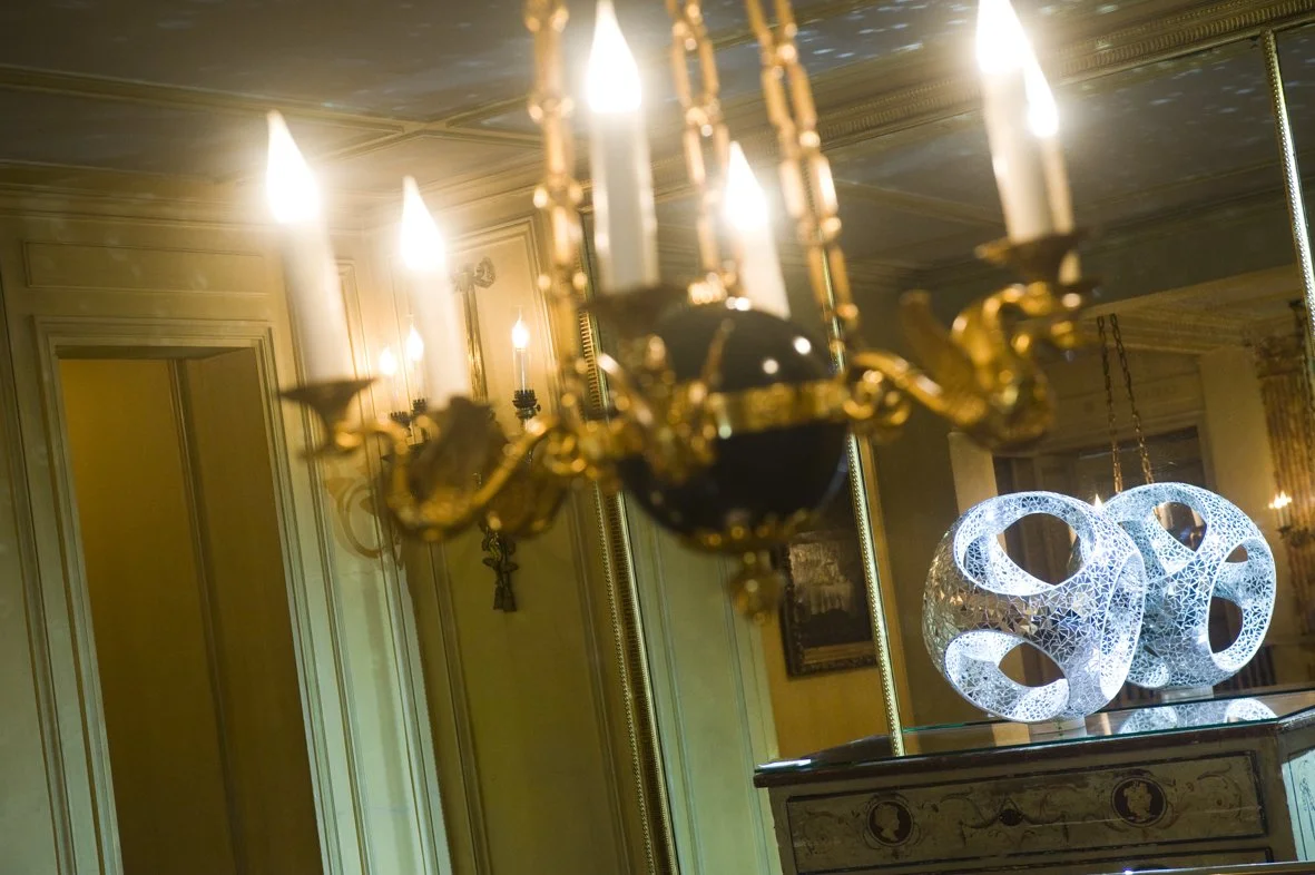 Un chandelier en or avec des bougies allumées, et deux sculptures modernes en forme de sphères illuminées avec des motifs géométriques dans un salon élégamment décoré.