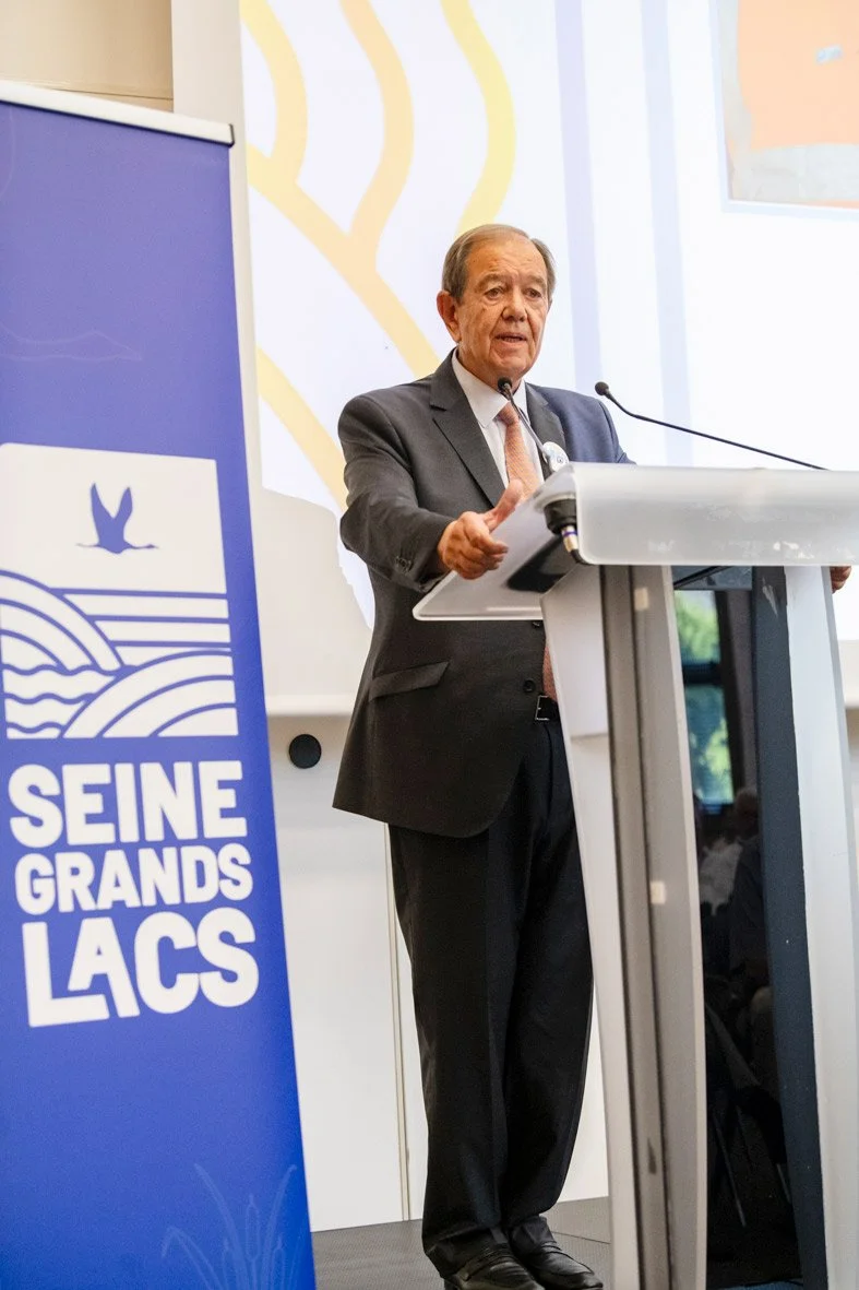 Homme en costume donnant une présentation ou un discours devant un micro lors d'un événement à l'intérieur. Logo de Seine-Grands-Lacs sur un panneau à côté.
