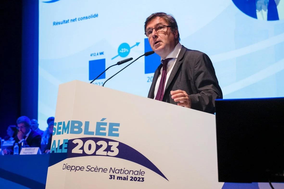 Un homme en costume noir et cravate, parlant lors d'une conférence, avec un graphique à l'écran en arrière-plan. Sur le pupitre, il y a une affiche indiquant 'SEMBLÉE ALE 2023', 'Dieppe Scène Nationale', et la date '31 mai 2023'.