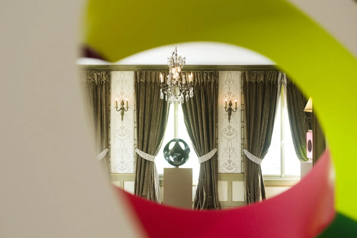 Une sculpture ronde et abstraite placée sur un socle au centre d'une salle élégante avec des lustres en cristal, des rideaux sombres et des murs ornés de motifs.