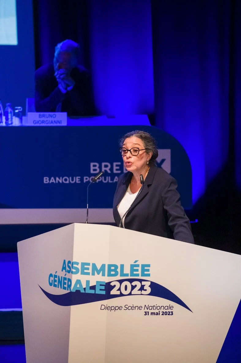 Femme parlant devant un podium lors de l'Assemblée Générale 2023 à Dieppe, portant des lunettes et une veste noire.