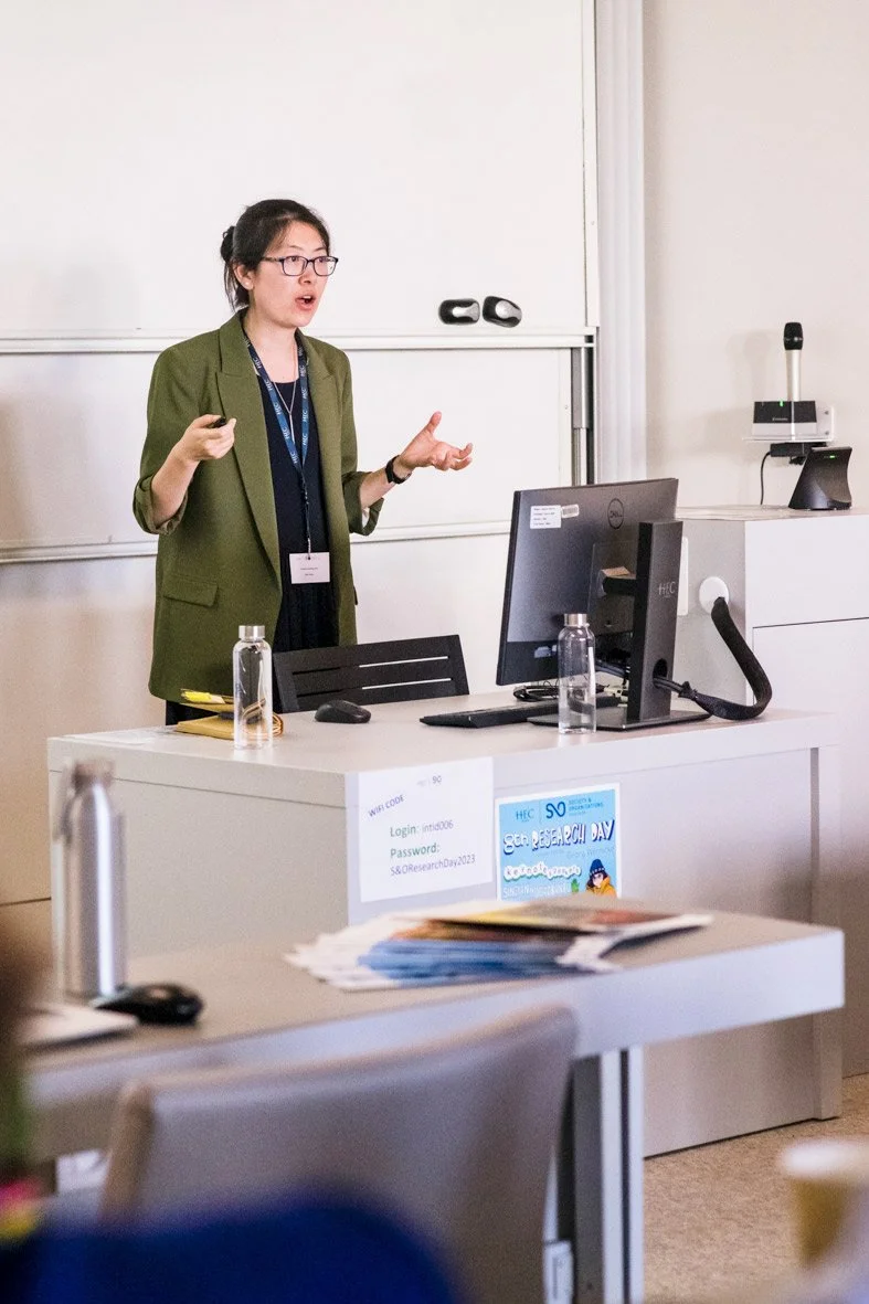 Une femme donnant une présentation dans une salle de classe ou de conférence, avec un tableau blanc derrière elle, une tablette devant elle, et des bouteilles d'eau sur le bureau.