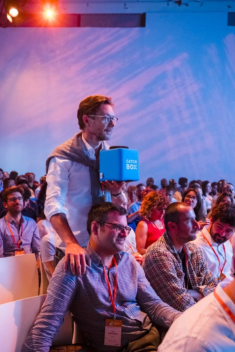 Un homme debout dans une salle de conférence, tenant une boîte bleue avec l'inscription "CATCH BOX", entouré de nombreux autres participants assis.