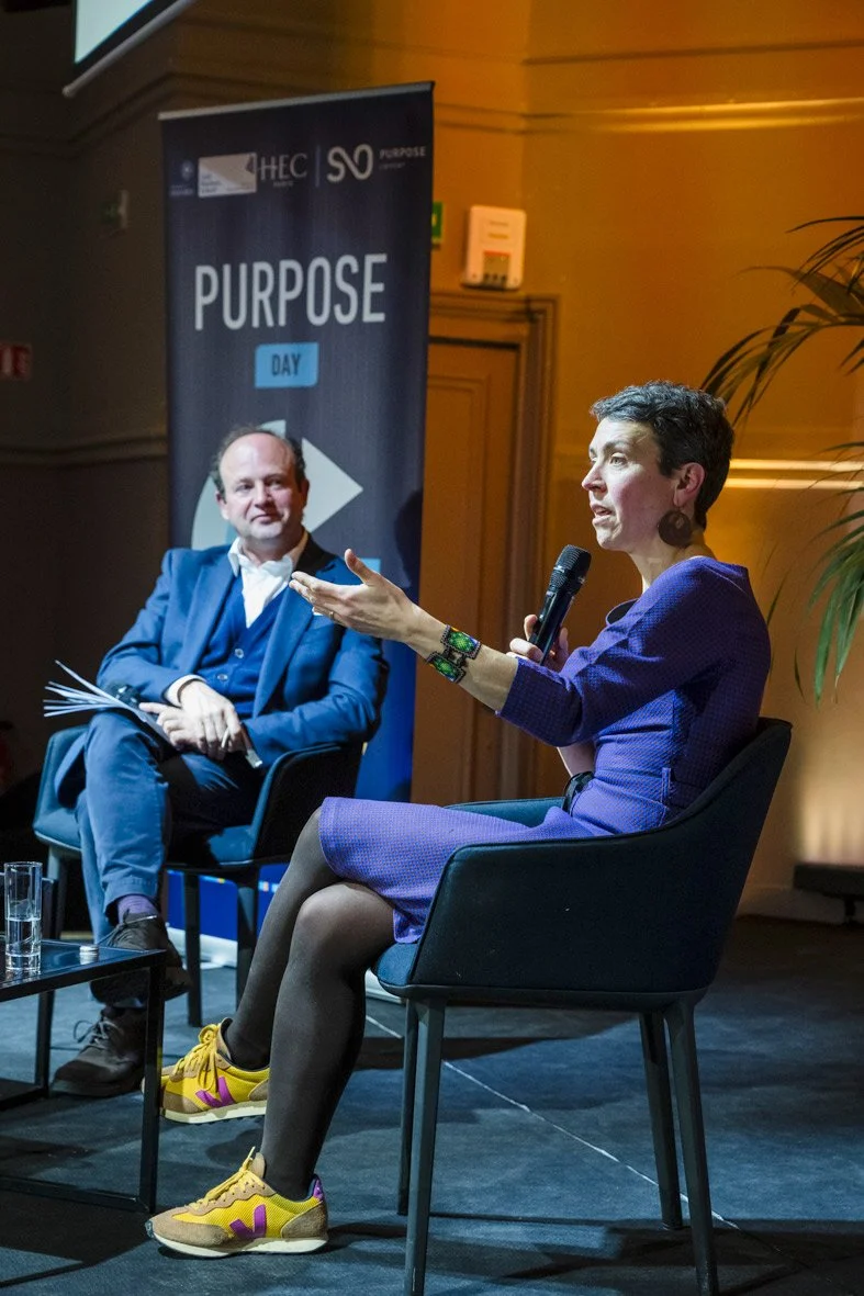 Une femme parle lors d'une conférence sur scène, accompagnée d'un homme assis. Une bannière derrière elle affiche 'PURPOSE DAY'.