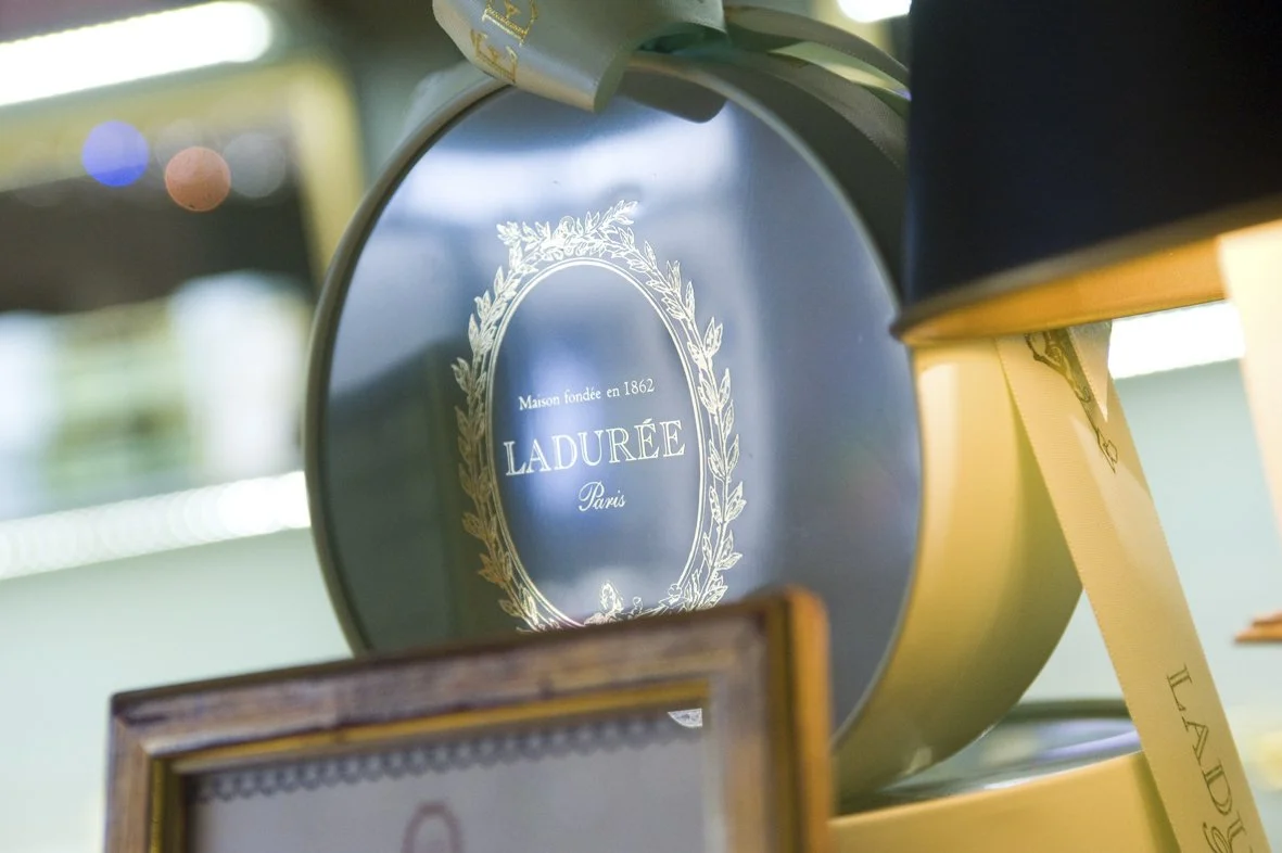 Boîte ronde de saveur dorée avec le texte 'Maison fondée en 1862 LADURÉE Paris' encadré d'une couronne de feuilles.