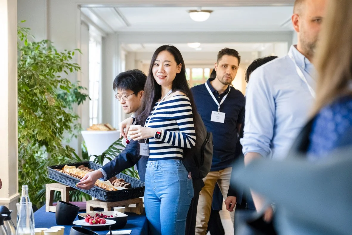 Des personnes lors d'un événement, une femme souriante sert des biscuits ou pâtisseries sur une table, d'autres personnes attendent ou se mêlent à proximité.