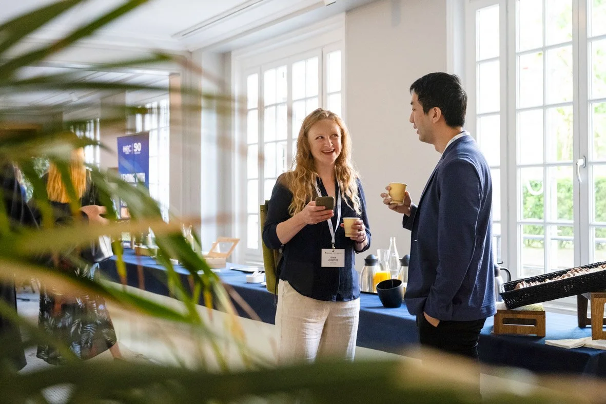 Deux personnes discutent lors d'une pause café dans une salle lumineuse avec de grandes fenêtres. La femme a les cheveux roux, porte un badge et tient une tasse, l'homme porte un costume et tient aussi une tasse.