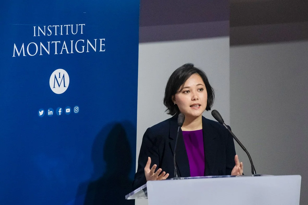 Femme parlant à un micro lors d'une conférence à l'Institut Montaigne, avec un panneau bleu à gauche affichant le nom de l'institut et des icônes de réseaux sociaux.