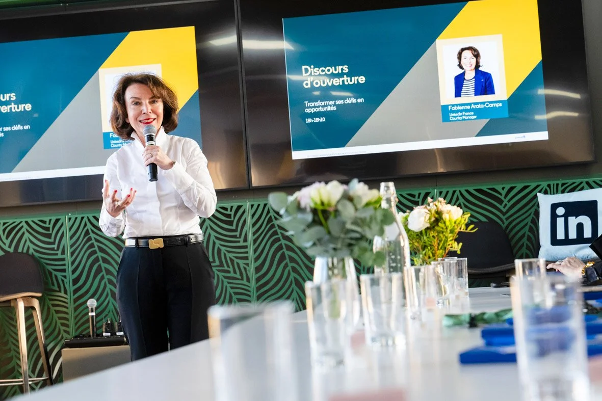 Une femme parlant lors d'une présentation dans une salle de réunion avec deux écrans et des fleurs sur la table.