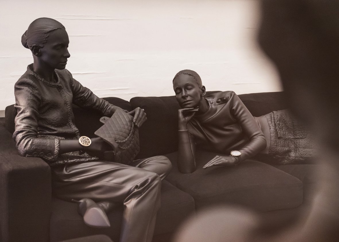 Sculptures de deux personnes en bronze, une femme et un homme, assises sur un canapé noir, dans un style réaliste.