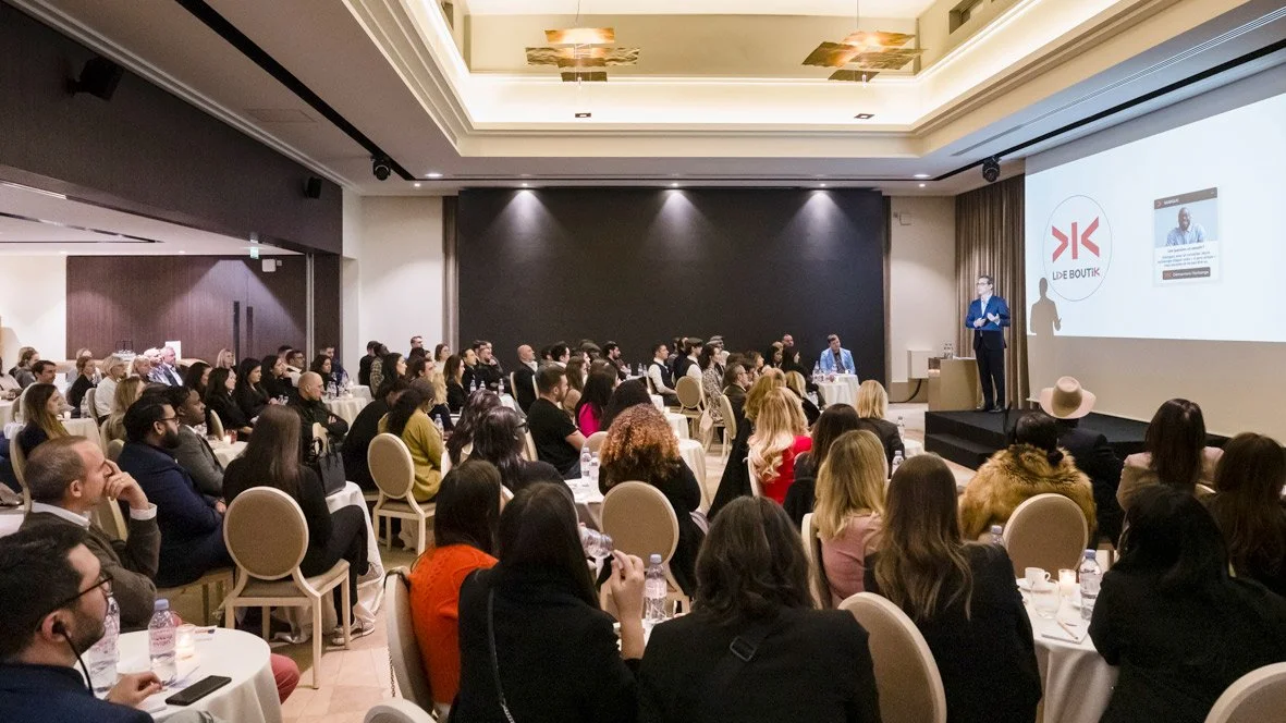 Conférence dans une salle avec un écran projecting une présentation et un orateur en costume sur scène, audience attentive.