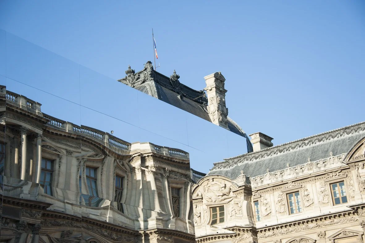 Reflet du toit d'un bâtiment historique dans une grande surface vitrée contre un ciel bleu clair.
