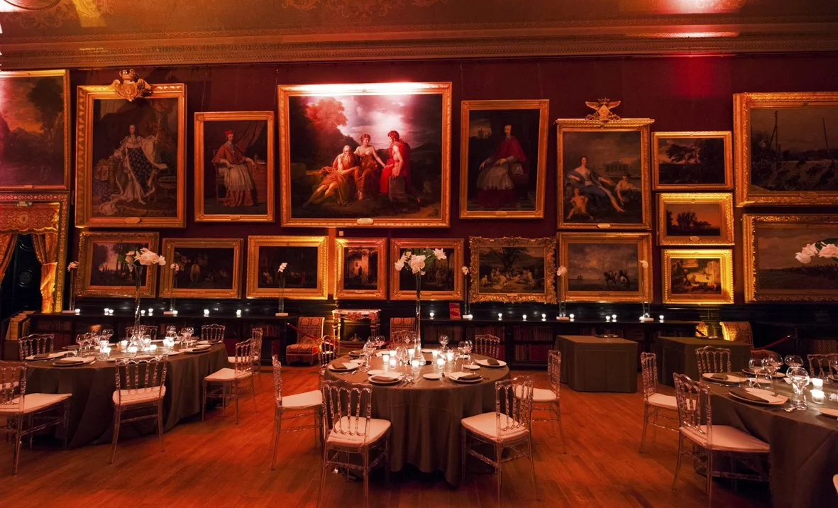 Une salle de réception ornée de nombreux tableaux anciens encadrés dorés, avec des tables rondes dressées pour un dîner élégant, et une ambiance chaleureuse avec un éclairage tamisé.