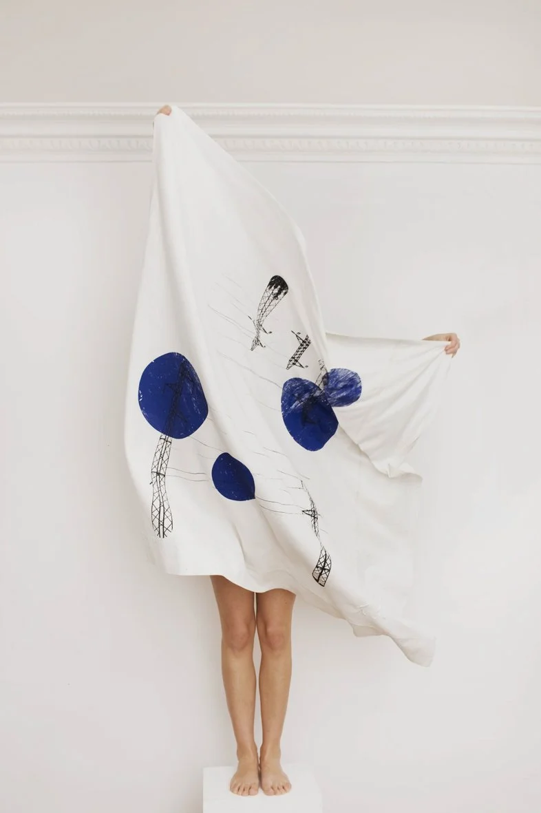 Personne debout derrière un drap blanc avec des dessins noirs et bleus de lampes et de ballons, tenant le drap sur les côtés.