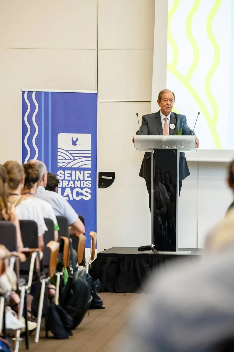 Homme en costume donnant une conférence devant un public dans une salle de réunion, avec un panneau indiquant 'Seine Grands Lacs' et un écran avec un fond jaune à lignes