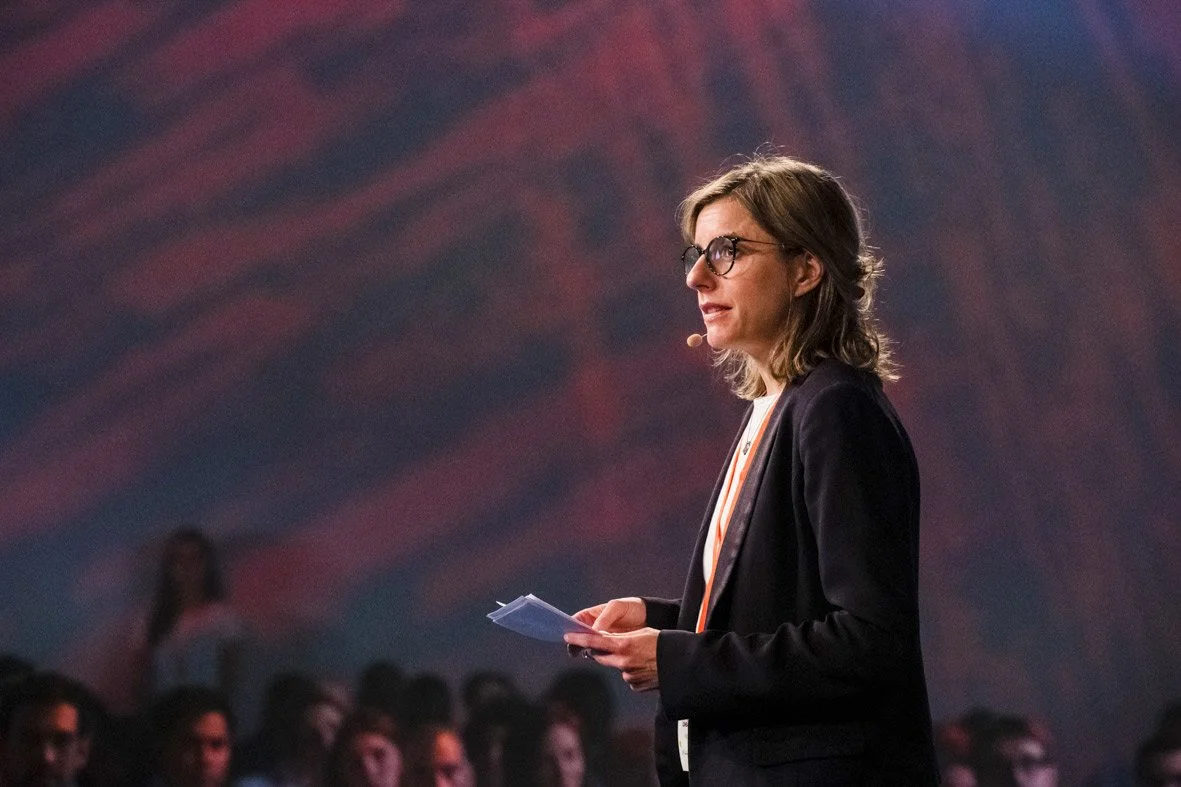 Une femme parlant lors d'une conférence, tenant des papiers, avec un micro attaché à l'oreille, dans une salle pleine d'auditeurs.