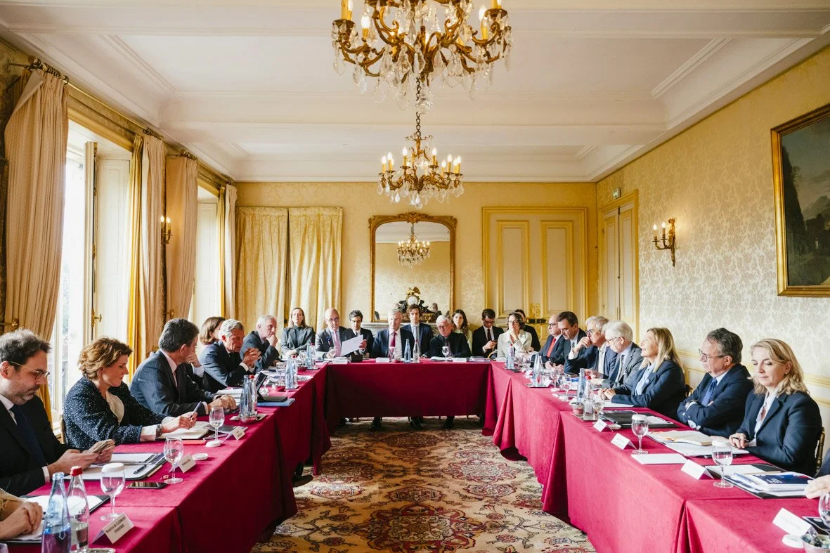 Réunion d'affaires avec de nombreux participants assis autour d'une grande table dans une salle élégante, décorée de chandeliers et de rideaux dorés.