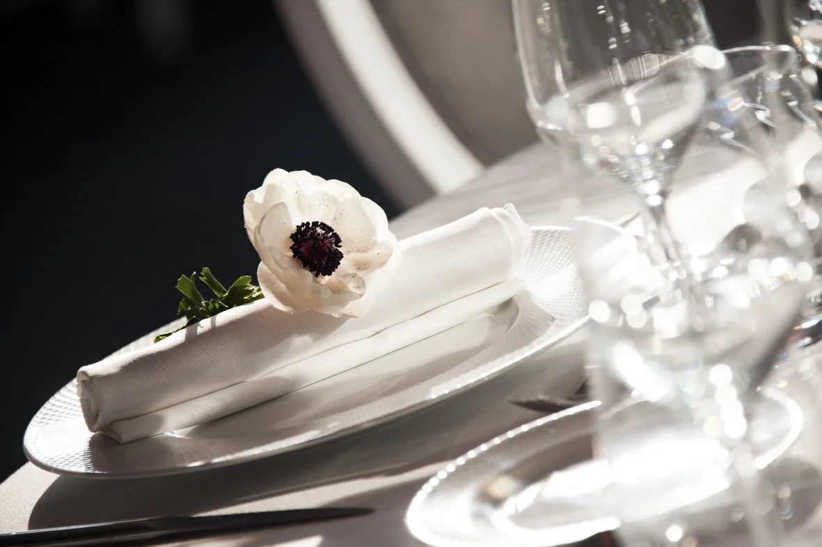 Une table de dîner élégante avec une nappe blanche, une assiette blanche, un porte-nom en tissu blanc avec une fleur blanche dessus, et plusieurs verres à vin en cristal transparents.