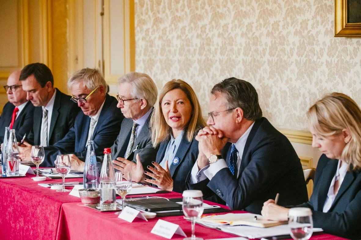 Un groupe de personnes assises à une table lors d'une réunion ou conférence, discutant et prenant des notes, avec des verres et bouteilles d'eau devant eux.